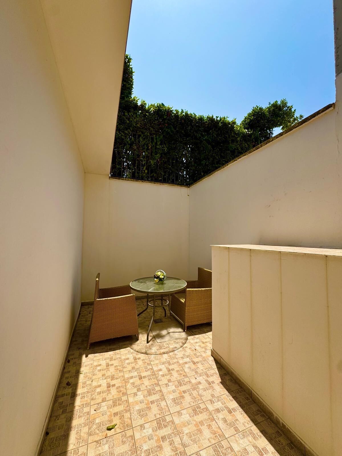 A small, enclosed patio area with a round table and wicker chairs, surrounded by tiled flooring and white walls.