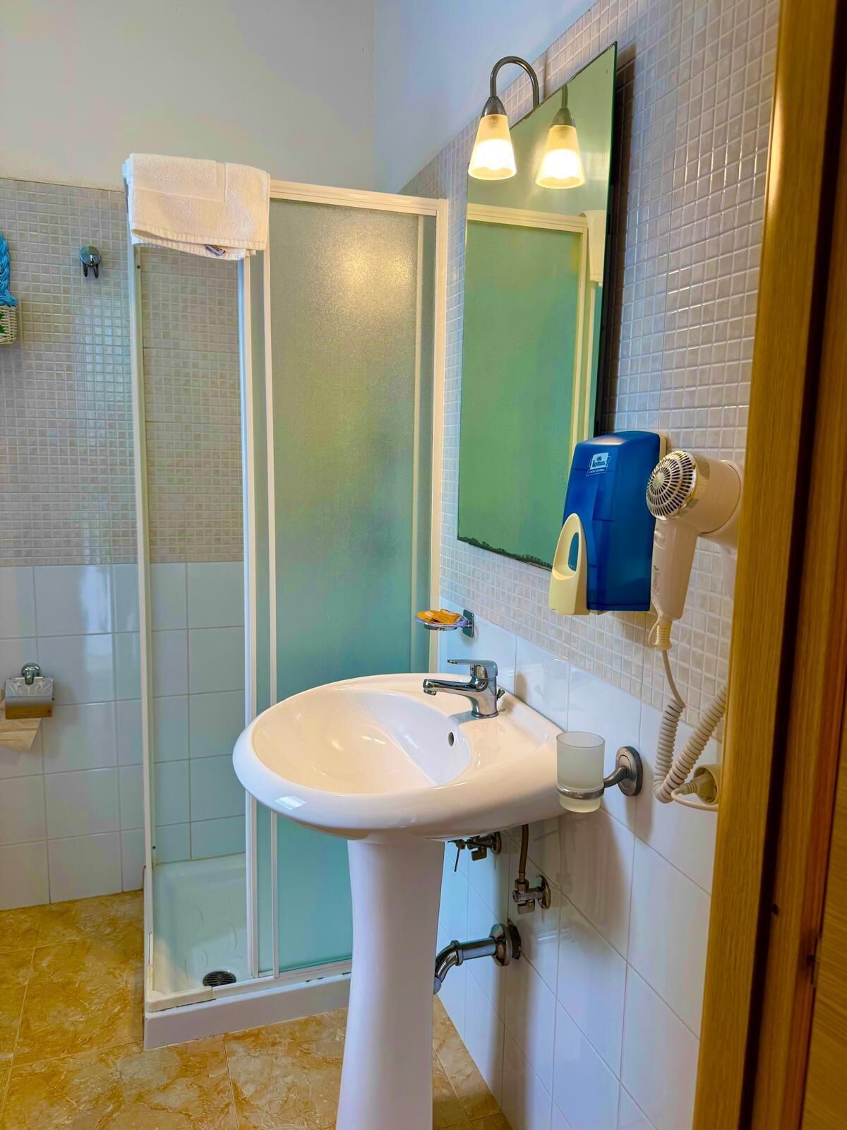 A bathroom with a white sink, a glass shower enclosure, a towel hanging on the door, and a mirror with a blue soap dispenser.