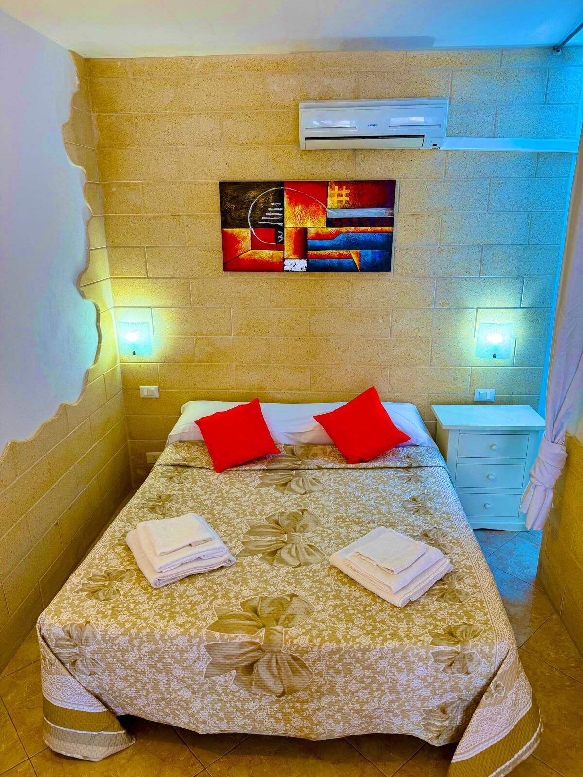 A hotel room with a bed, two red pillows, a nightstand, an air conditioning unit, and a wall-mounted artwork.