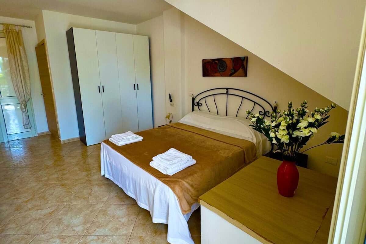 A bedroom with a bed, a wooden nightstand, a red vase with flowers, a white wardrobe, and a painting on the wall.