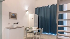 A small kitchenette with a toaster, a kettle, and a cup on the counter. There is a chair and a table in front of a window with blue curtains.