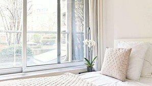 Un dormitorio con un amplio ventanal, una cama con ropa de cama blanca y una planta en maceta.