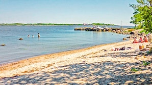 Ein sandiger Strand mit badenden und entspannten Menschen. Boote liegen im Wasser vor Anker und ein Steg führt ins Meer.