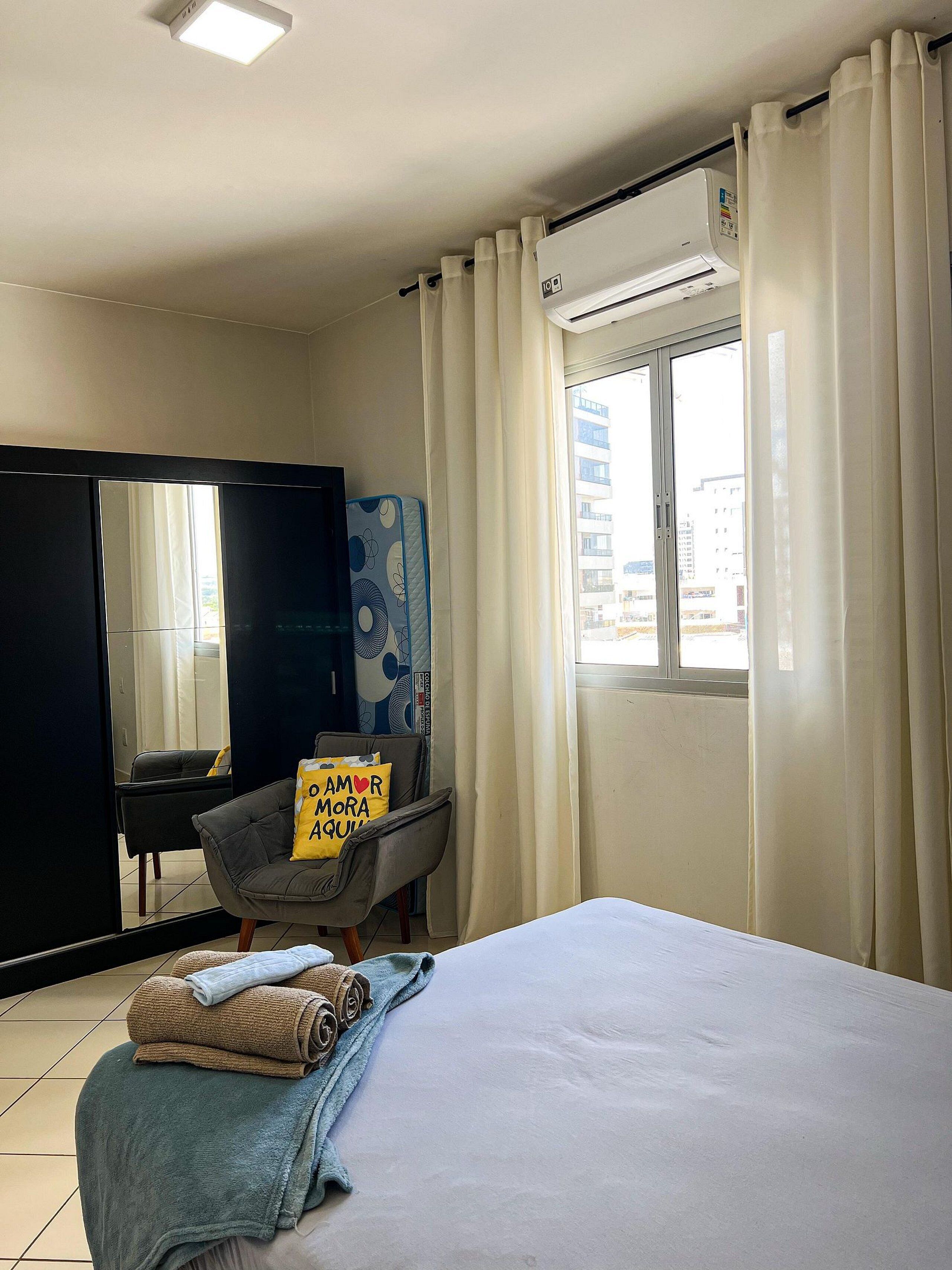 A bedroom with a bed, a window with curtains, a wall-mounted air conditioner, a mirror, and a chair with a pillow.