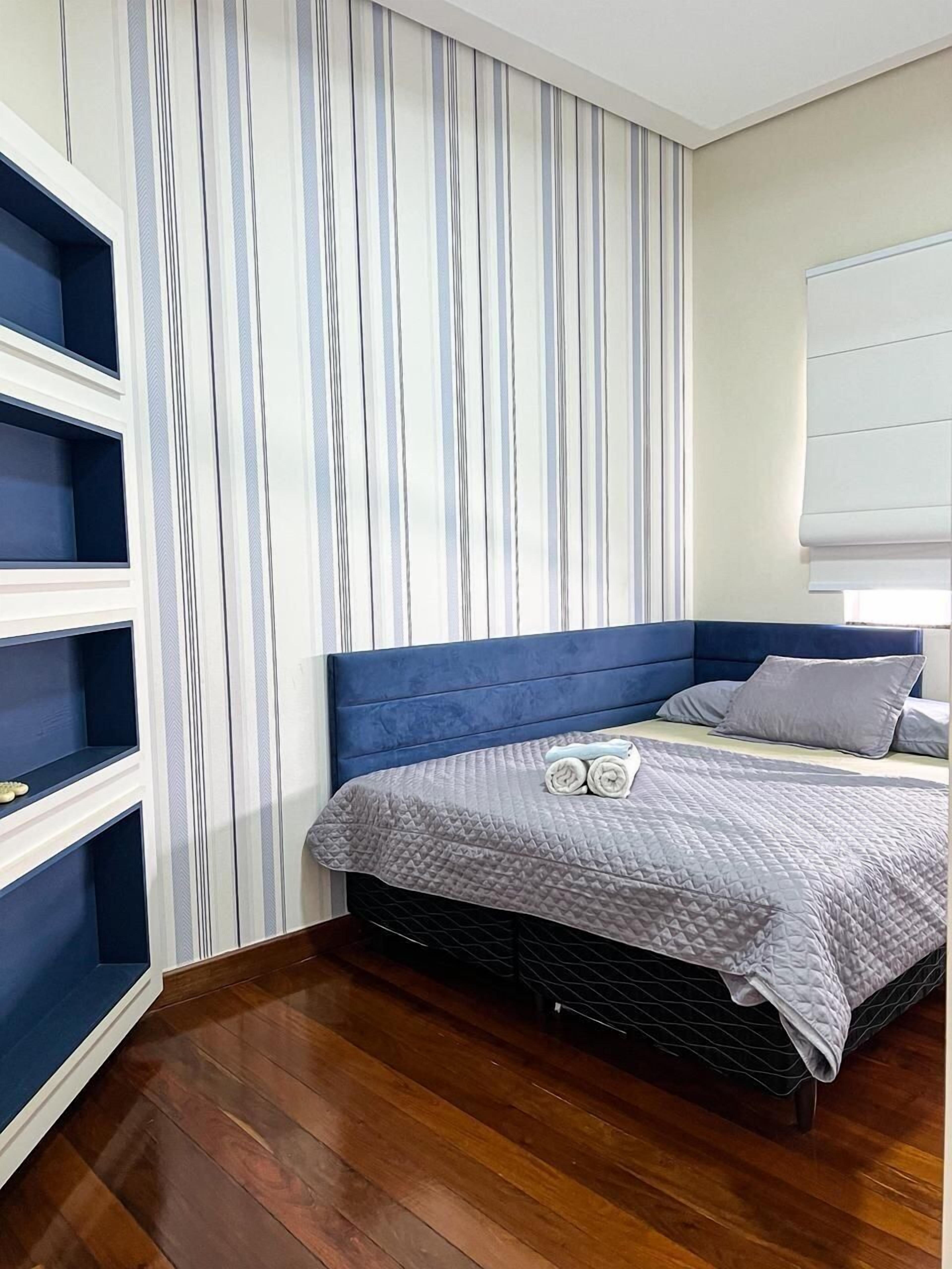 A bedroom with a blue headboard, a grey quilted bedspread, and a striped curtain.