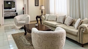 A modern living room with a sectional sofa, two armchairs, a coffee table, and a rug. There is a large window with curtains, a ceiling light, and a framed picture on the wall.