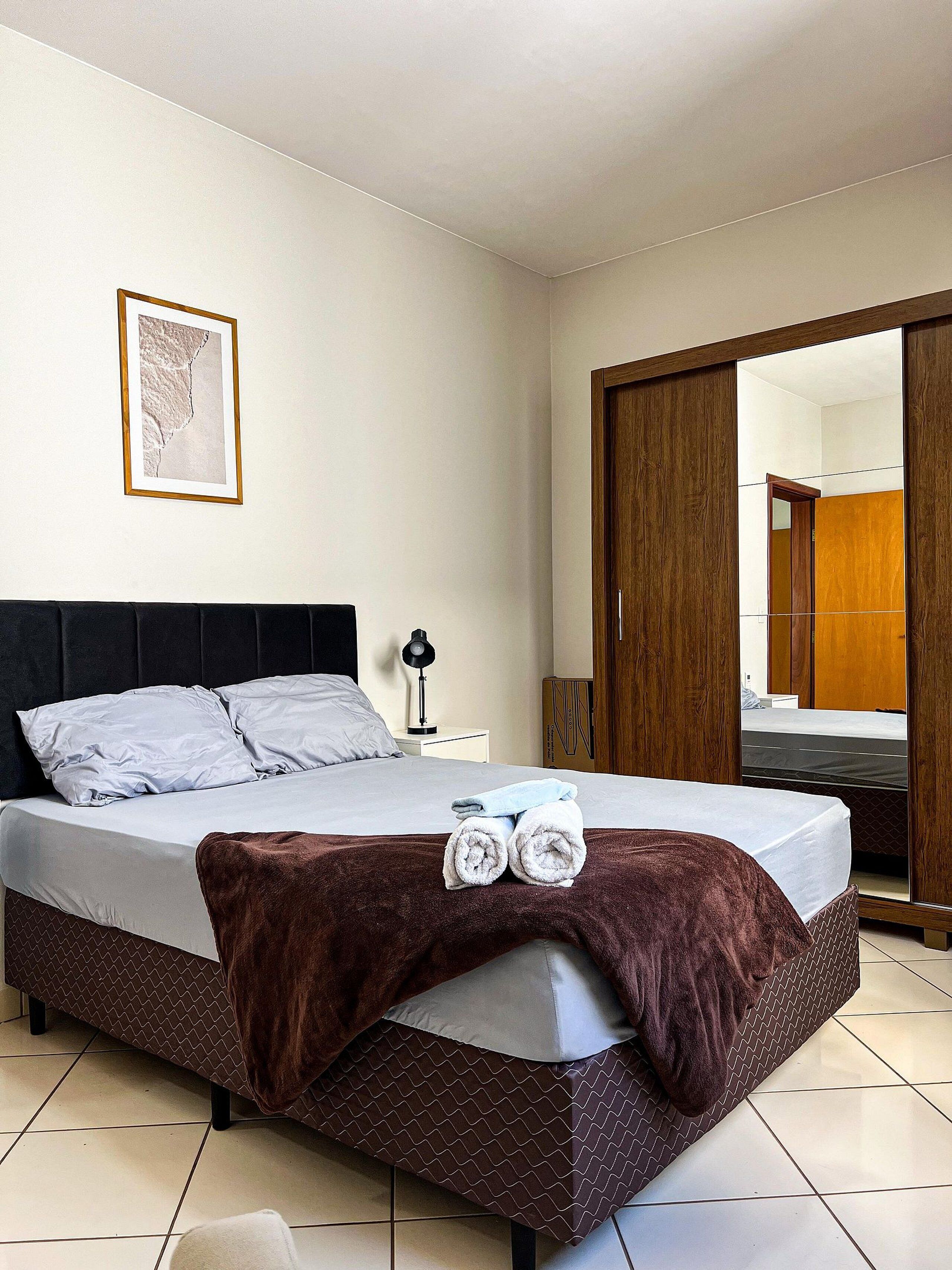A neatly made bed with a brown throw blanket and two rolled towels, a bedside lamp, a framed picture on the wall, and a large mirror reflecting a wardrobe and a bed.