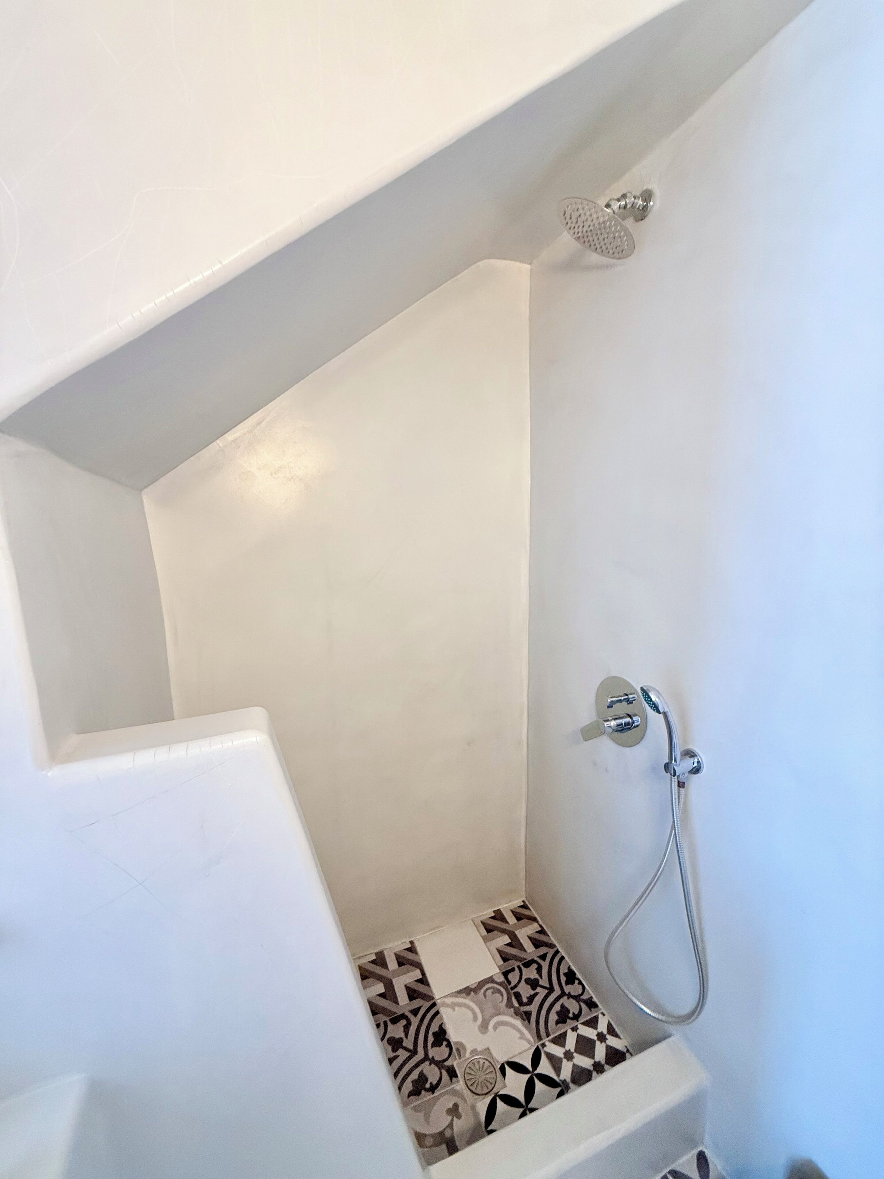 A small, enclosed shower area with a showerhead and hose, tiled floor with a geometric pattern, and white walls.