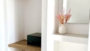 Un coin de pièce avec une étagère en bois, un panier tressé et un miroir sur lequel est posé un vase de fleurs séchées.