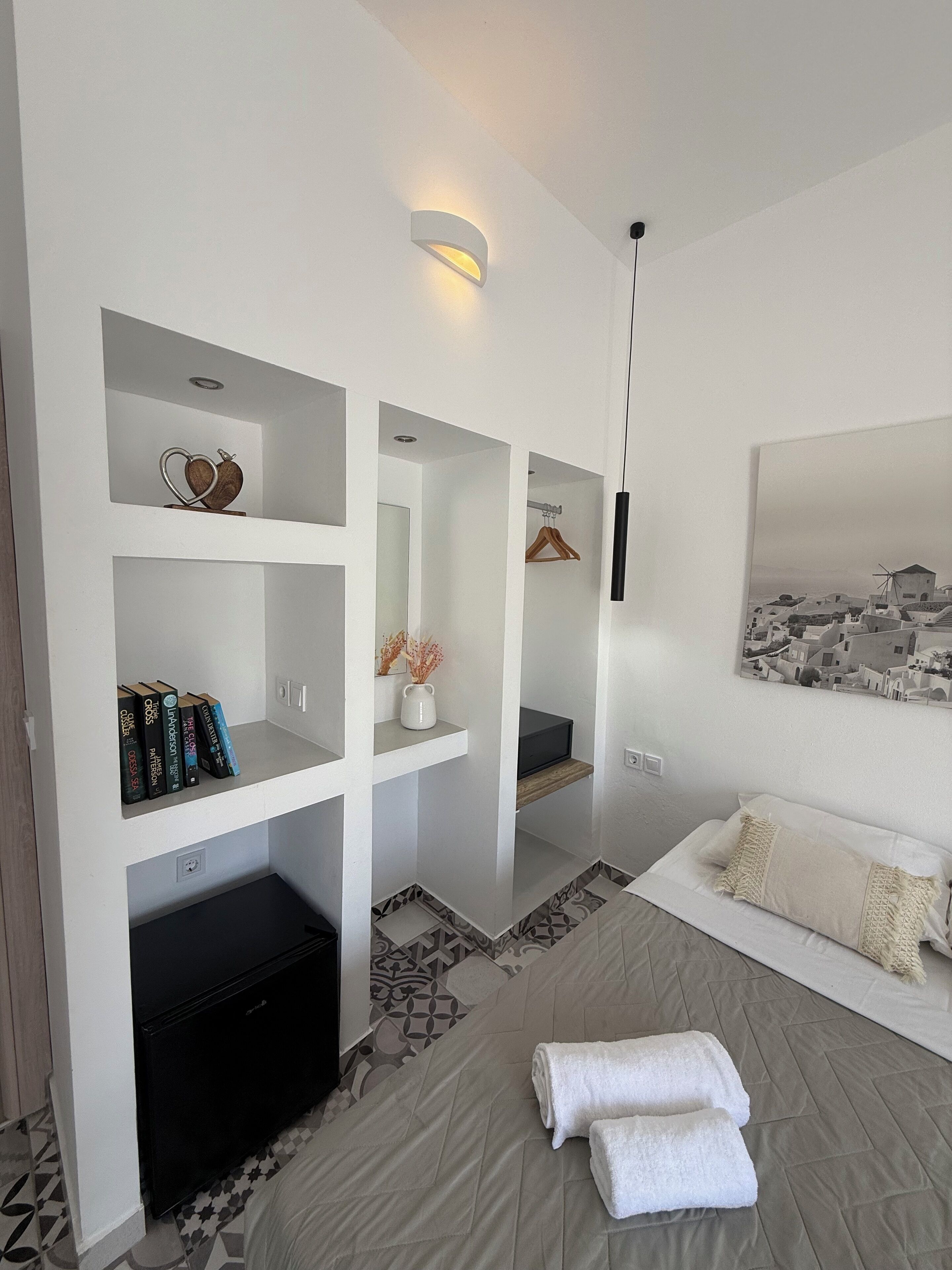 A modern hotel room with a bed, a TV, a bookshelf, and a painting of a coastal village.