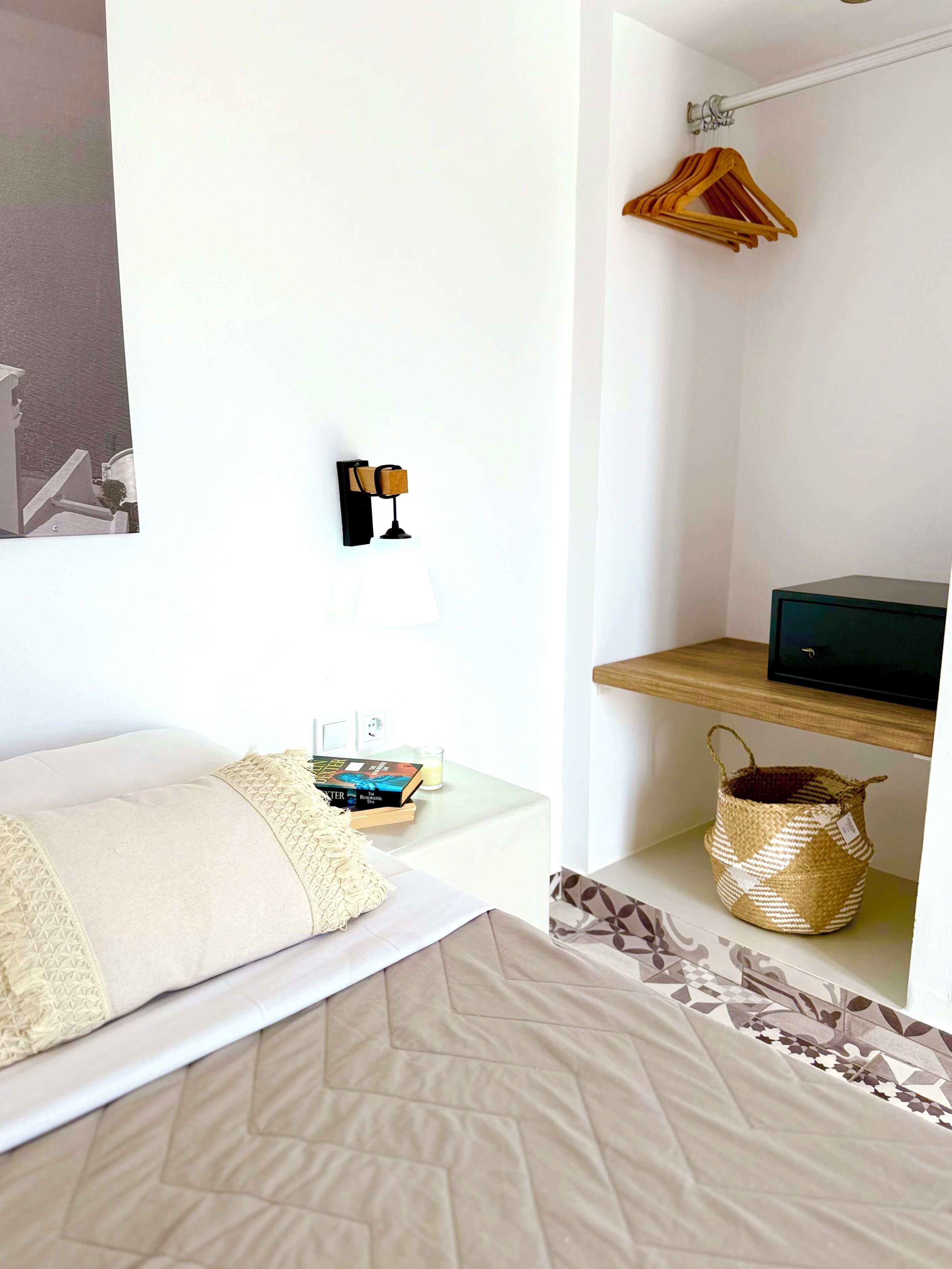 Habitación de hotel ordenada con cama, mesita de noche, lámpara y una pequeña estantería con una cesta.