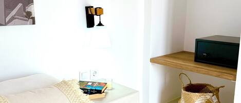 Habitación de hotel ordenada con cama, mesita de noche, lámpara y una pequeña estantería con una cesta.