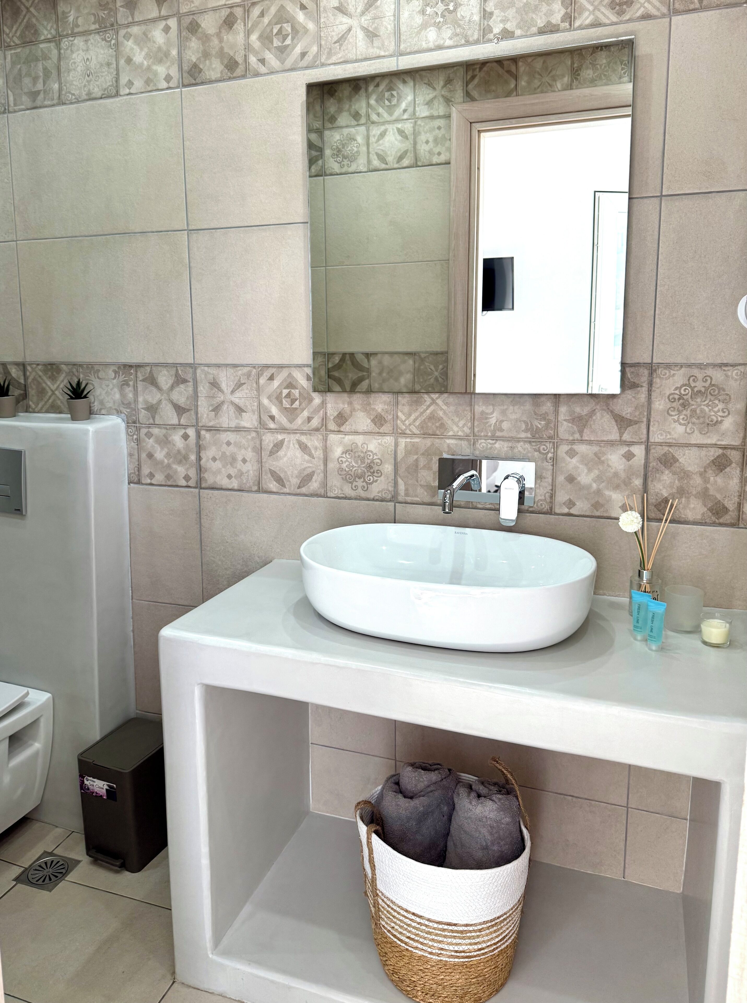 Baño con un lavamanos blanco, un espejo encima y una pared de azulejos con un diseño geométrico. Hay un cubo de basura y una cesta con toallas debajo del lavamanos.