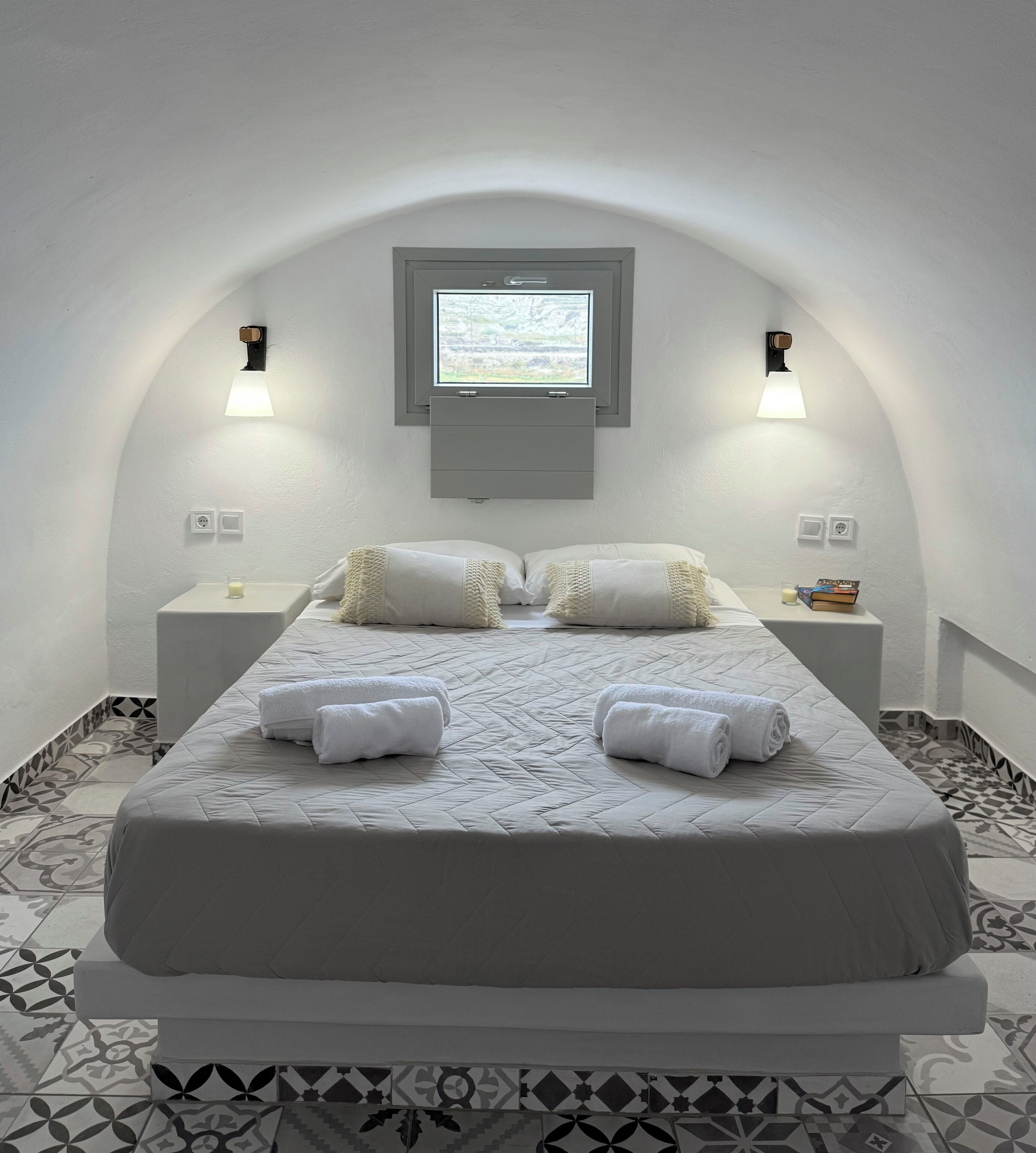 Un dormitorio con una cama, dos mesitas de noche, una ventana y una lámpara fijada a la pared.