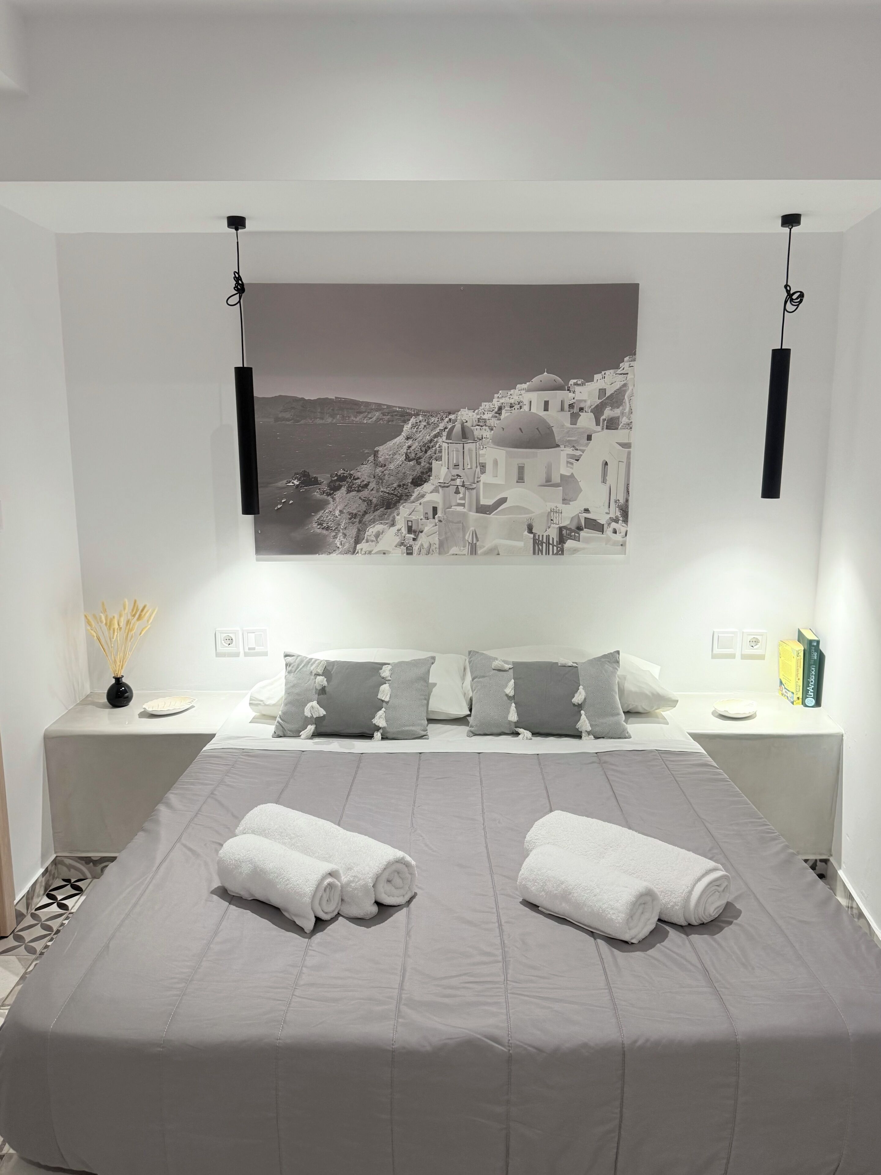 A modern hotel room with a large bed, two bedside tables, a wall-mounted picture, and a view of a coastal town.