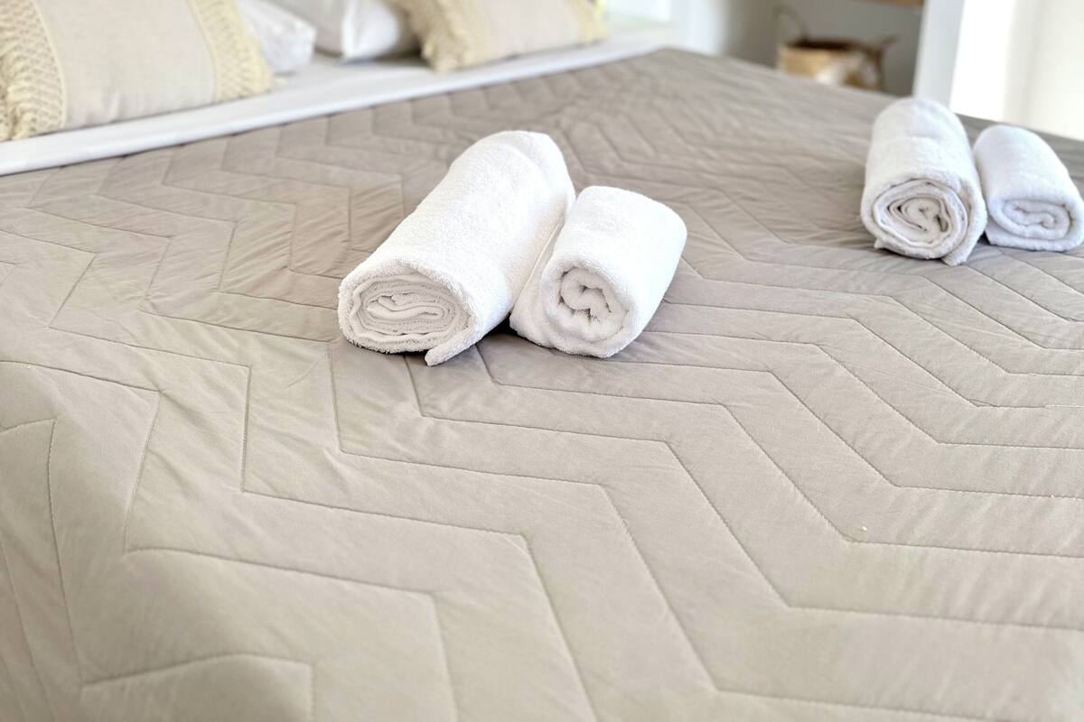 Una cama bien hecha con toallas blancas enrolladas, un cabecero acolchado moderno y una decoración minimalista con una fotografía blanco y negro enmarcada en la pared.