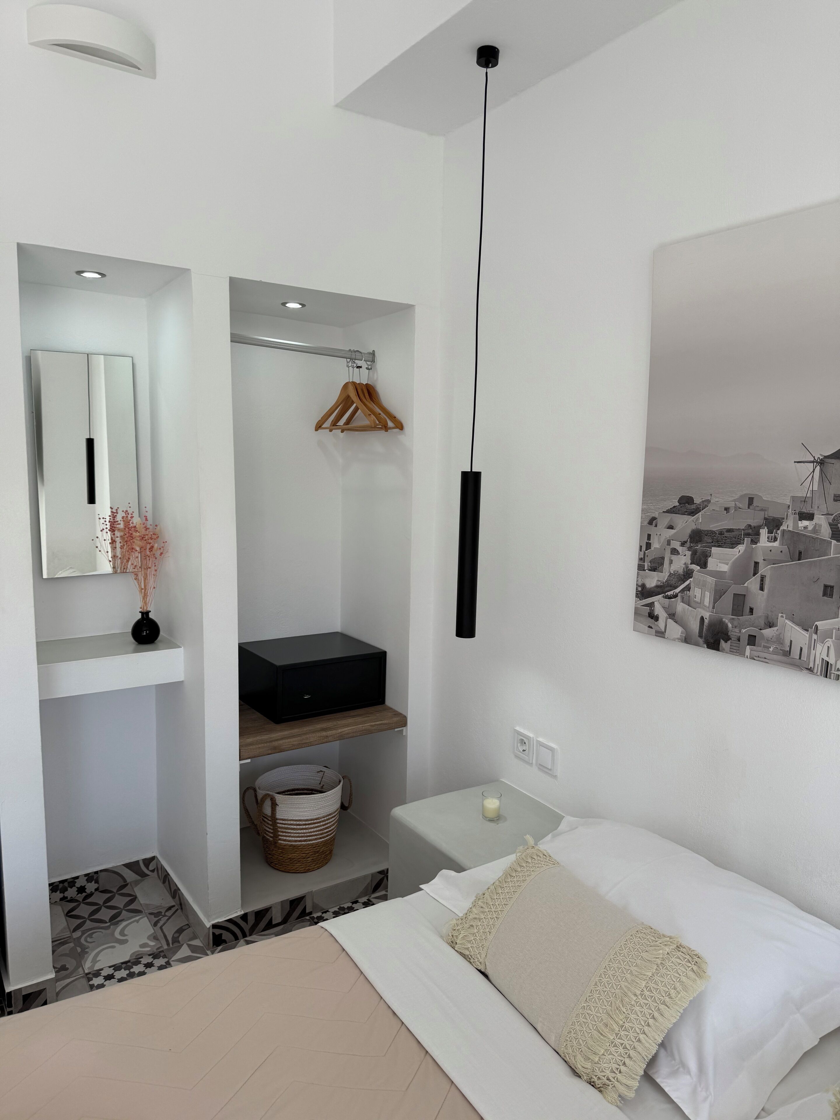 Un dormitorio moderno con una cama, una mesita de noche con lámpara, un espejo, un mueble negro para guardado, una percha para ropa y un cuadro colgado en la pared de un pueblo costero.