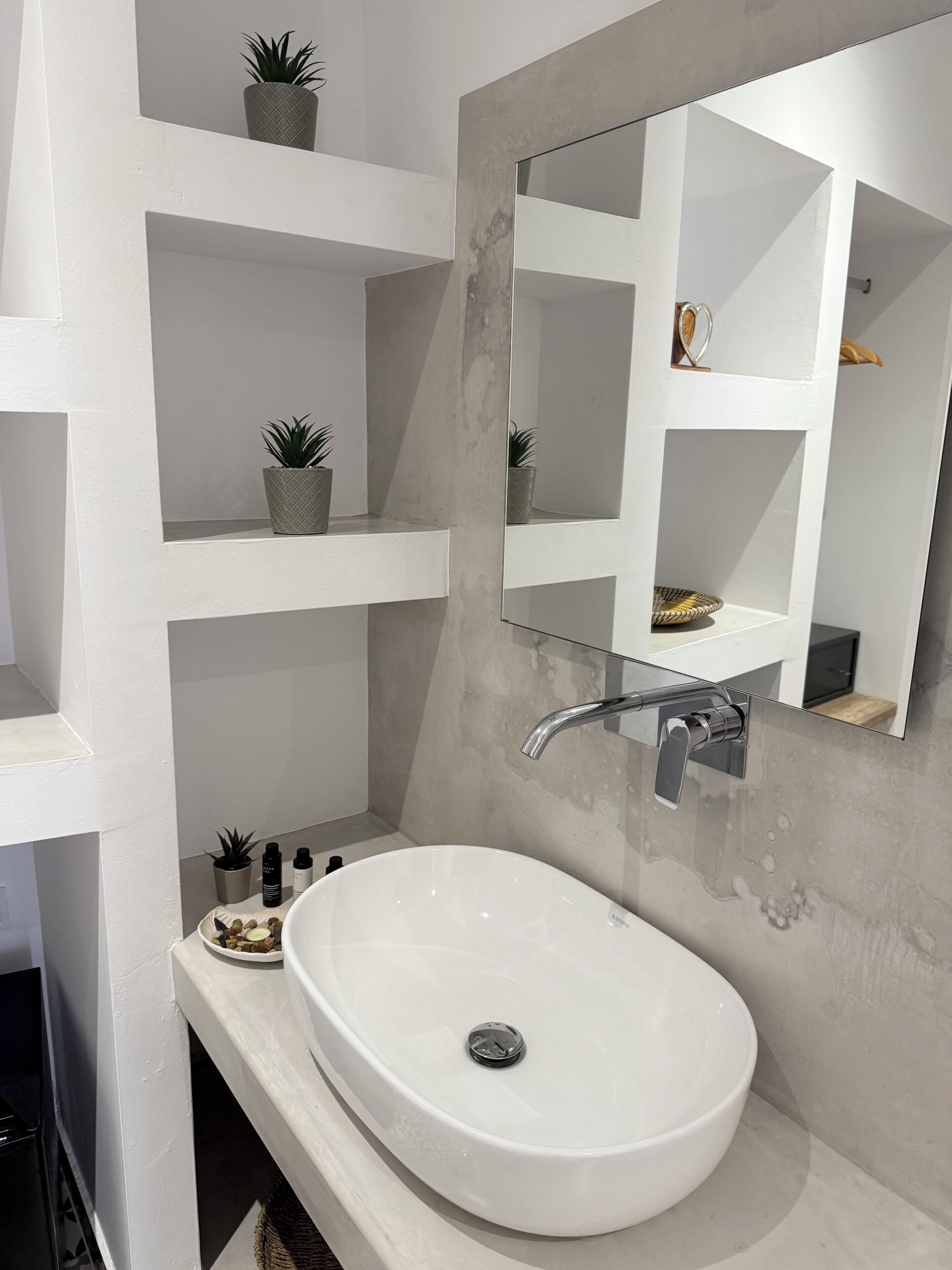 A modern bathroom with a white sink, a large mirror, and shelves with potted plants.