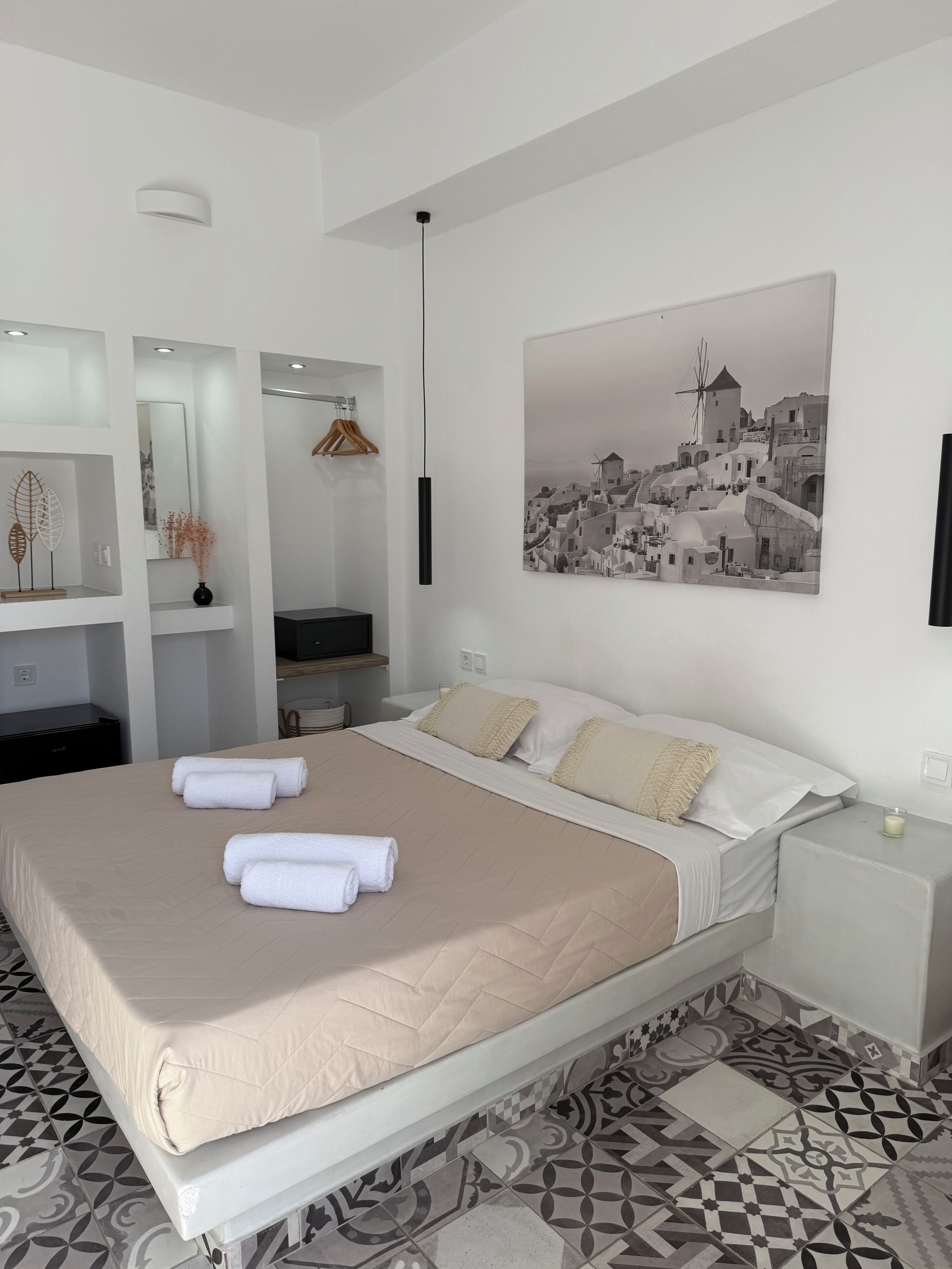 A modern hotel room with a large bed, a bedside table with a candle, a wall-mounted TV, and a painting of a coastal town.