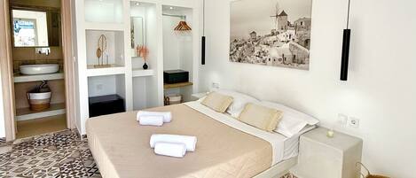 Un dormitorio moderno con una cama grande, una mesita de noche, un televisor montado en la pared y un baño con lavabo y espejo.