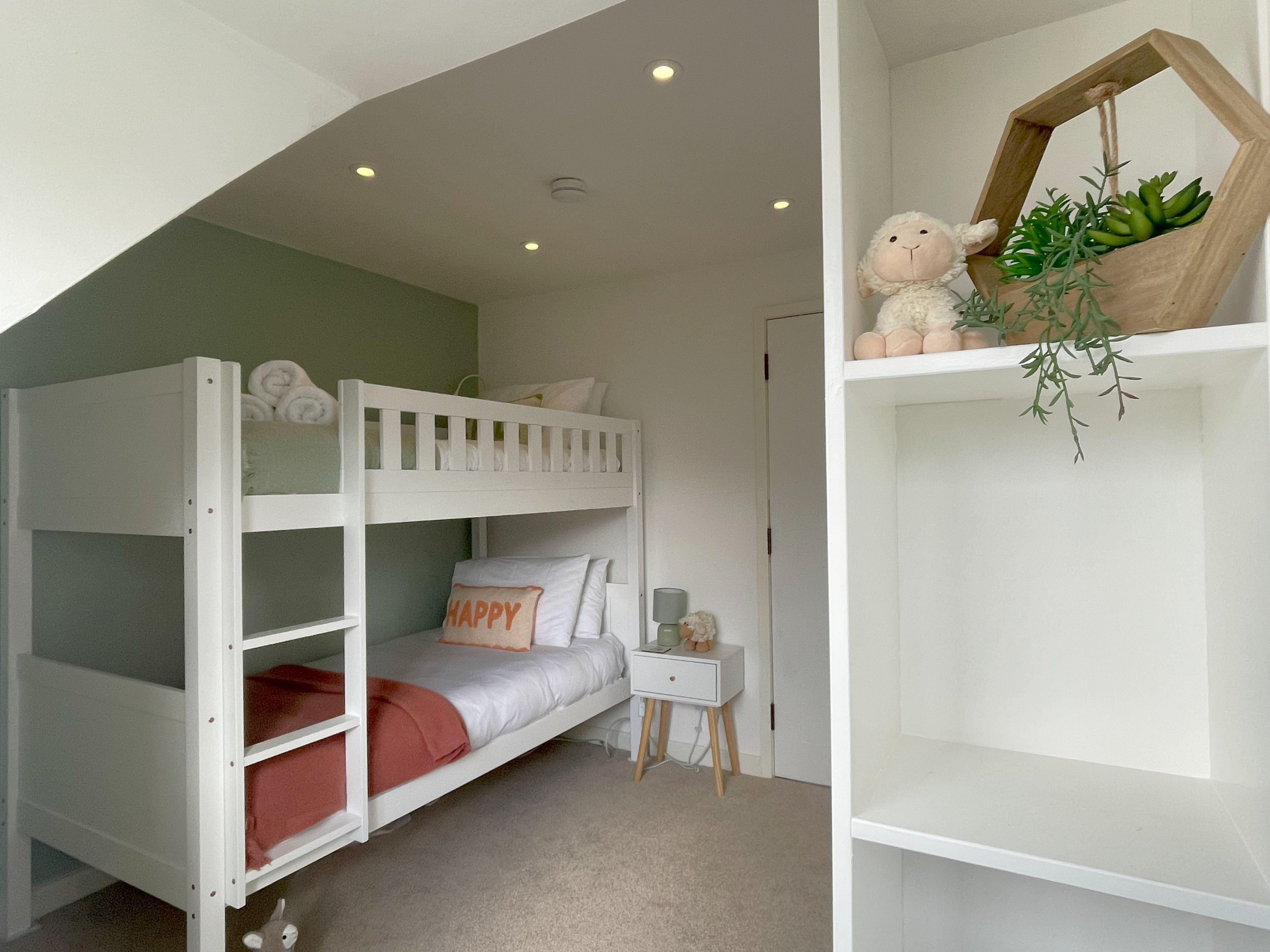 A bunk bed with a 'Happy' pillow, a small bedside table with a lamp, and a shelf with a stuffed animal and a plant.