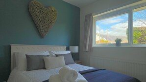 Ein ordentlich bezogenes Bett mit weißen Leinentüchern und zusammengerollten Handtüchern, eine herzförmige Wanddekoration und ein Fenster mit Blick ins Freie.