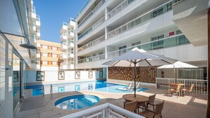 Un área de piscina en la azotea con sillones y sombrillas, rodeada por un edificio de varios pisos con balcones.