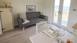 Ein gemütliches Wohnzimmer mit grauem Sofa, einem Esstisch mit Weinglas und Blick durch eine Glastür.