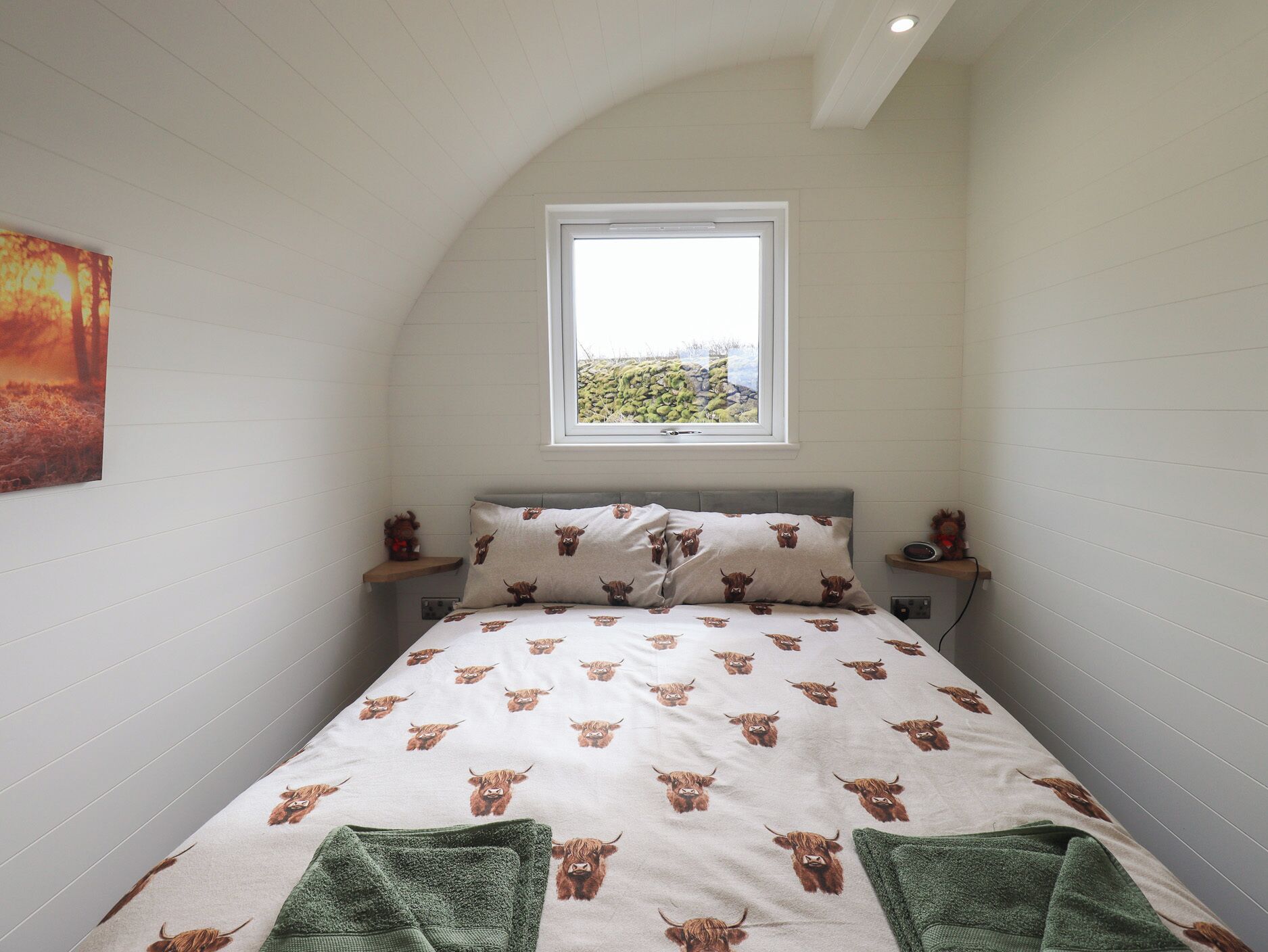 Un dormitorio con techo inclinado, una cama con edredón blanco estampado con motivos de animales marrones, dos toallas verdes, una mesita de noche con un radio y una ventana con vistas a la vegetación.