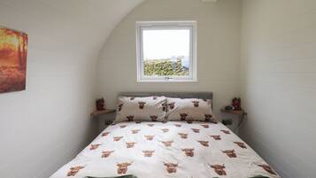 Un dormitorio con techo inclinado, una cama con edredón blanco estampado con motivos de animales marrones, dos toallas verdes, una mesita de noche con un radio y una ventana con vistas a la vegetación.