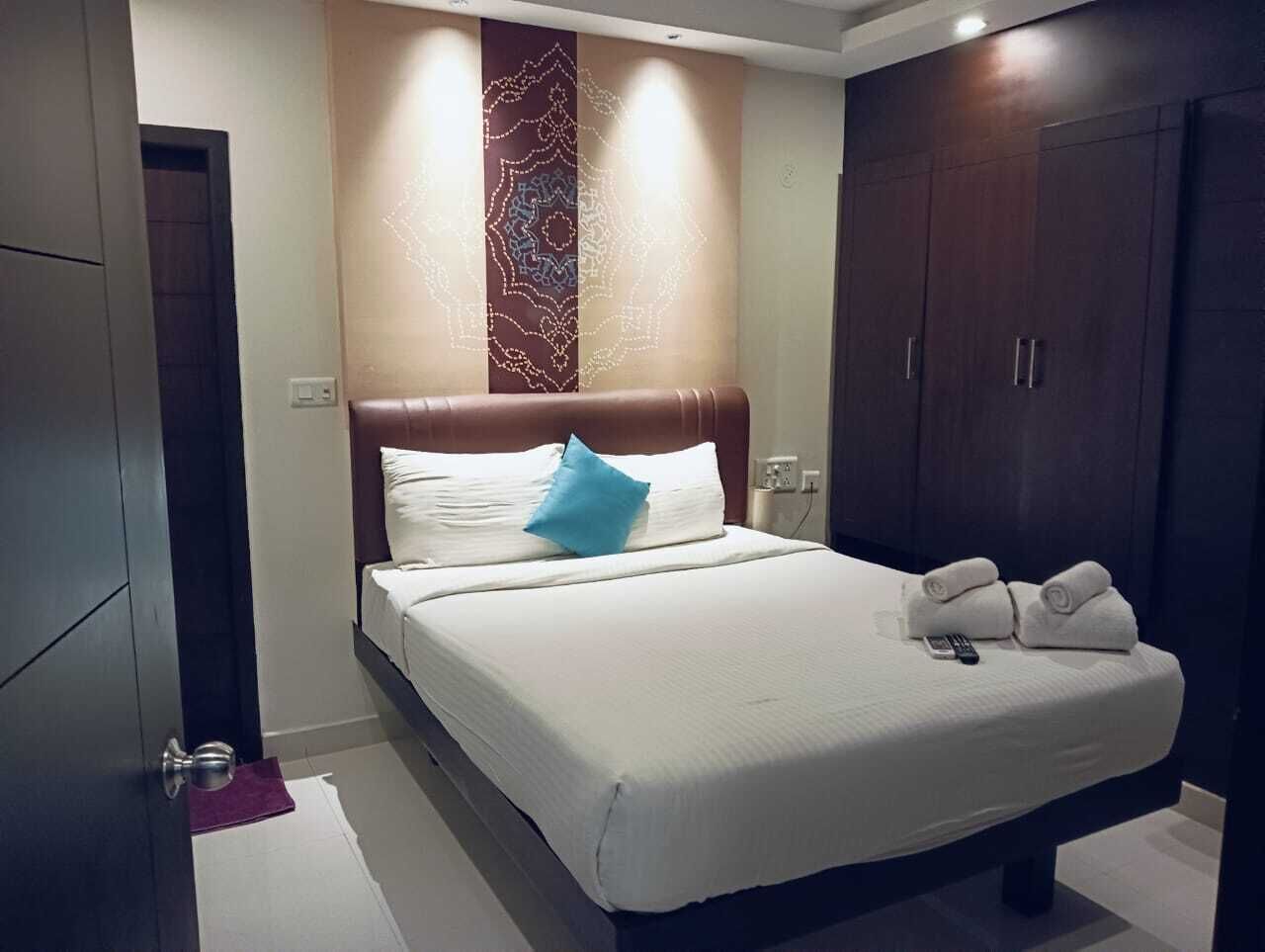 Una habitación de hotel con una cama grande, cabecera con panel decorativo, armario y mesita de noche con mando a distancia y toallas.