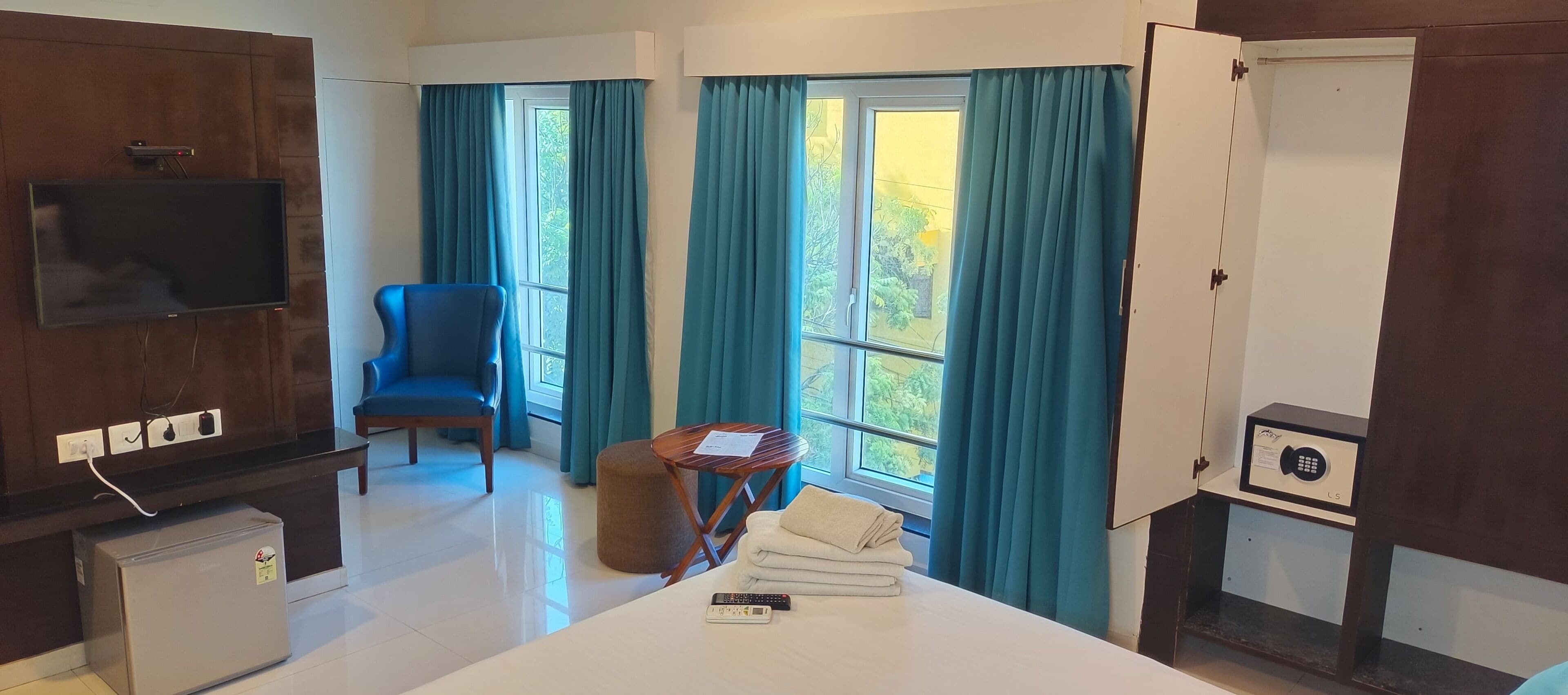 A hotel room with a bed, a chair, a small table, a TV, a minibar, and a window with curtains.