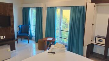 A hotel room with a bed, a chair, a small table, a TV, a minibar, and a window with curtains.
