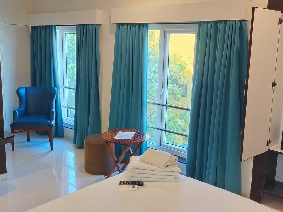 A hotel room with a bed, a chair, a small table, a TV, a minibar, and a window with curtains.