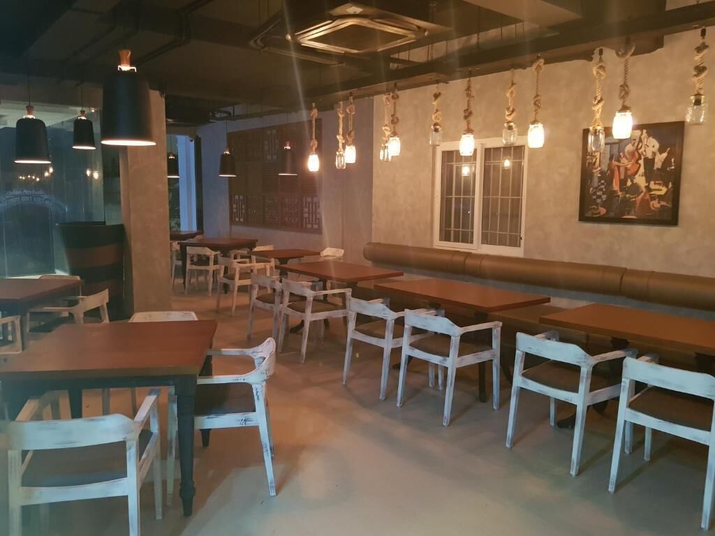 A restaurant interior with wooden tables and chairs, hanging pendant lights, and a large window.