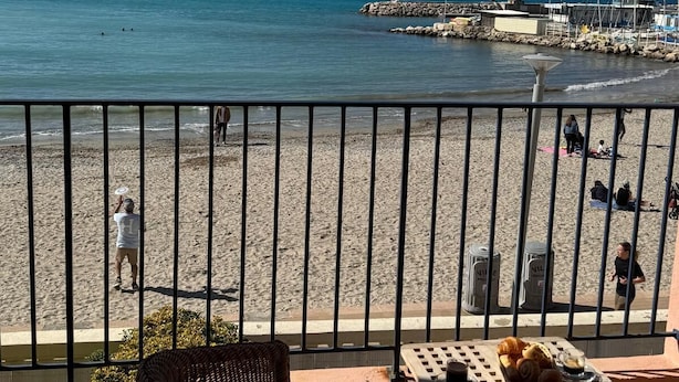 Un balcón con sillas de mimbre y una mesa con pasteles y una taza, con vistas a una playa con gente y barcos.