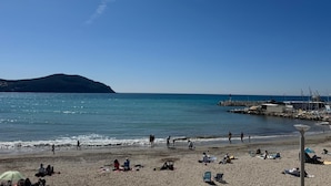 Una playa con gente tomando el sol y caminando por la orilla, un muelle con barcos y un cielo despejado.