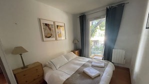 Un dormitorio con cama, mesita de noche, lámpara, ventana con cortinas y dos cuadros en la pared.