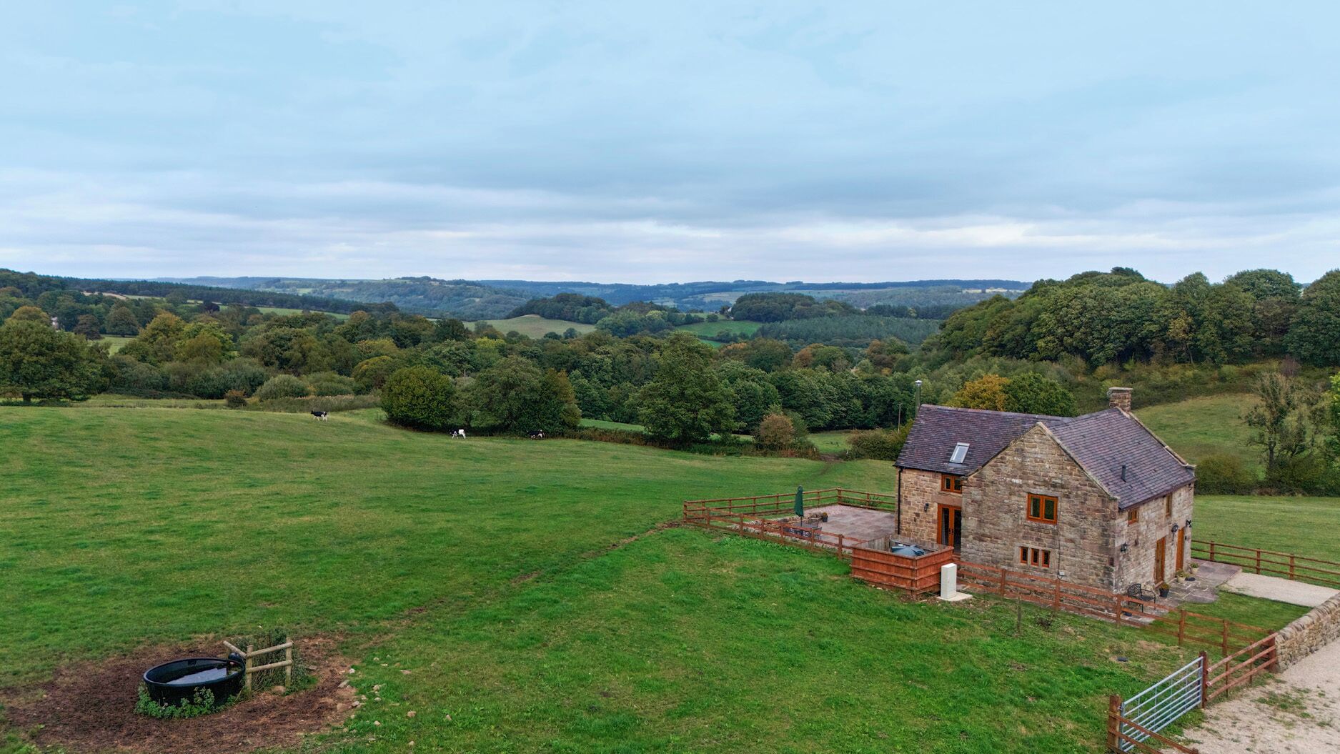 Une ferme en pierre avec une cour clôturée, un petit espace extérieur aménagé pour s’asseoir et une vue sur des collines verdoyantes et des arbres au loin.