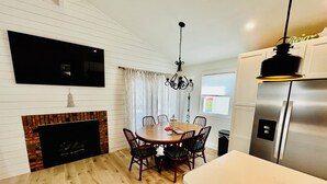 A dining area with a wooden table and chairs, a fireplace, a flat-screen TV mounted on the wall, and a modern refrigerator.