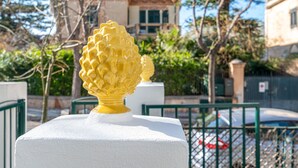A white pedestal with a yellow decorative object on top, set against a backdrop of a building and greenery. - Mondello Sea Life Duplex by Wonderful Italy (Palermo)