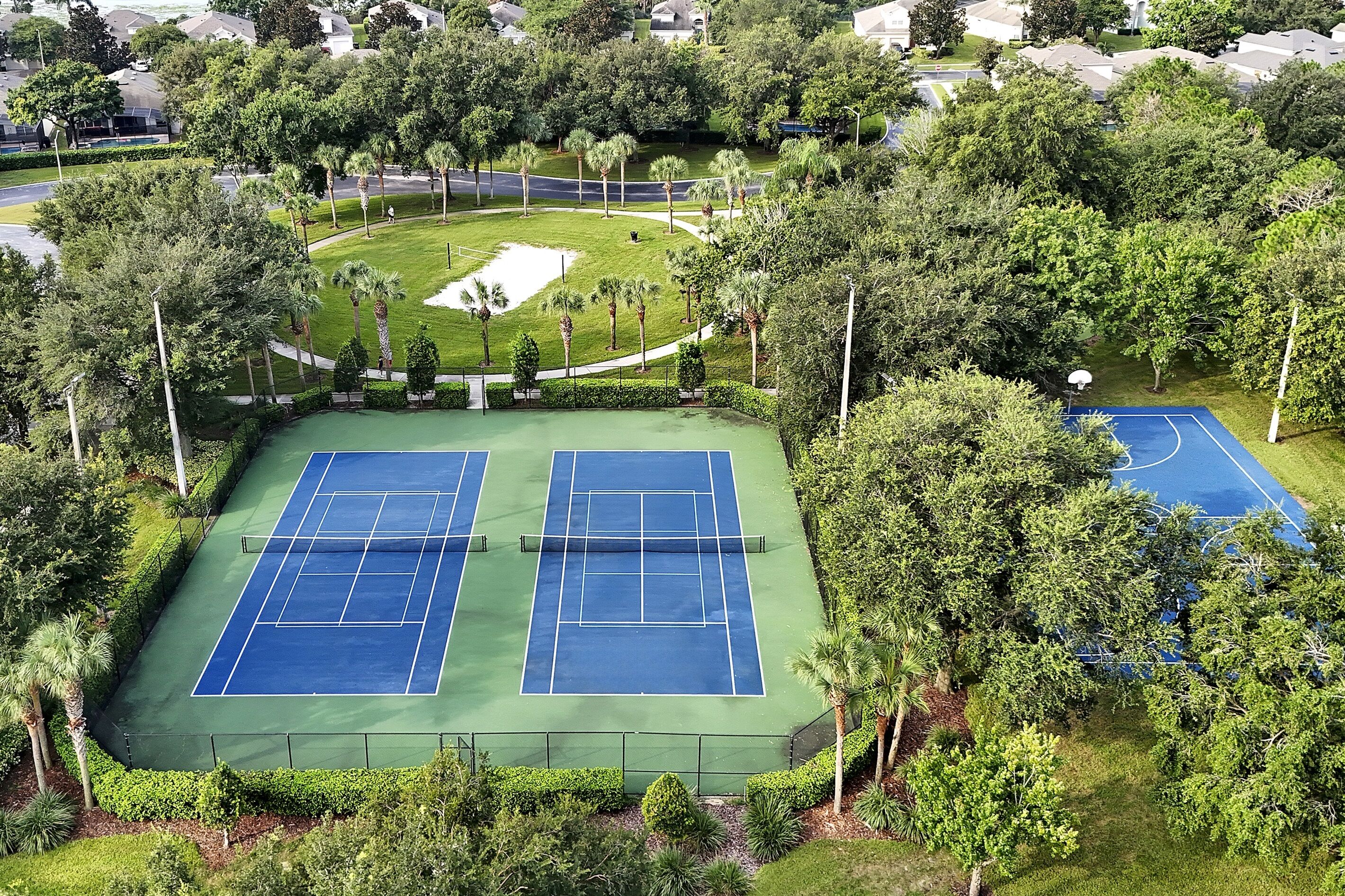 Vue aérienne d’un complexe de courts de tennis, entouré d’une végétation luxuriante et de bâtiments résidentiels.