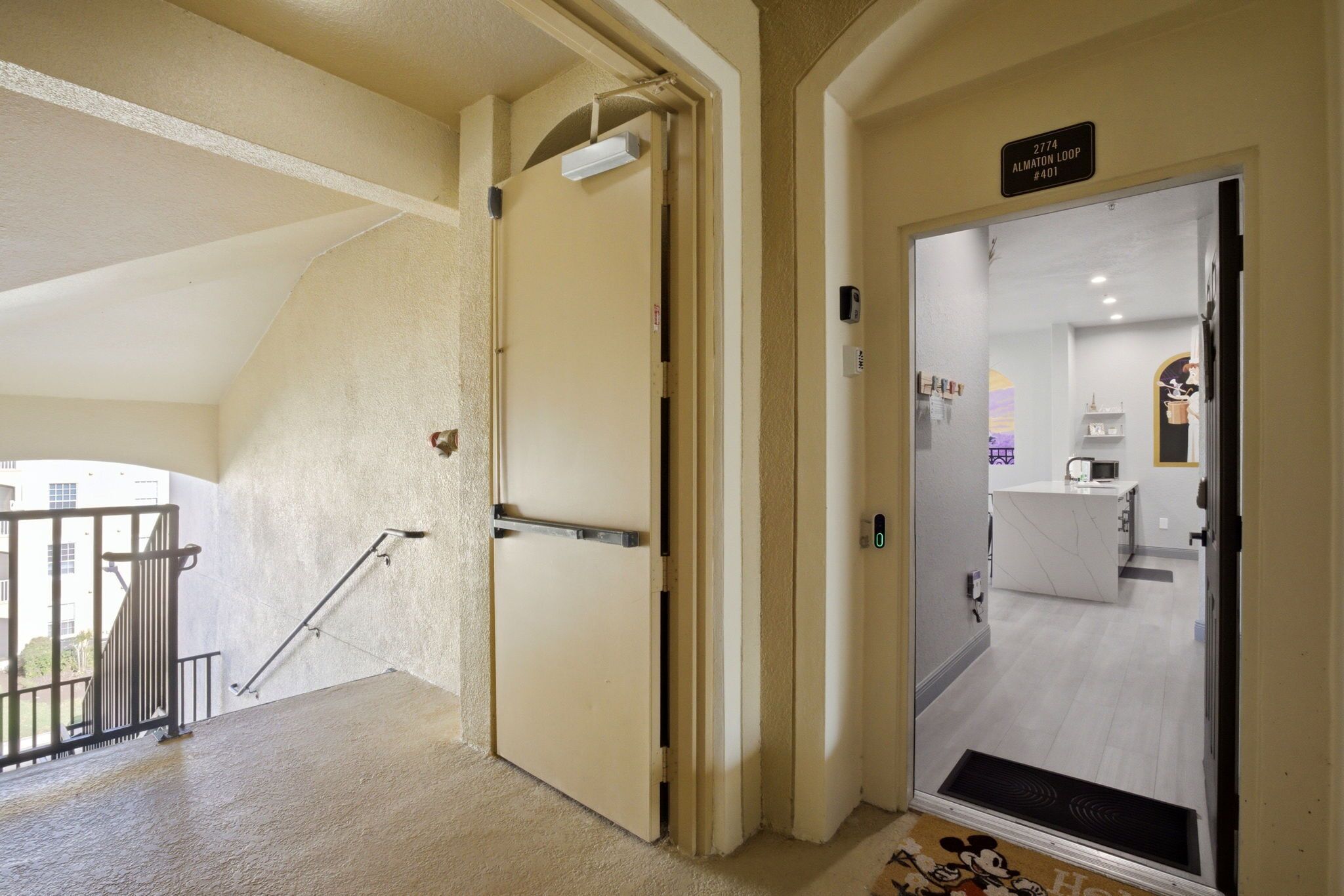 Entrada de um hotel com uma porta identificada como '2774 Almaton Loop #401', uma pequena cozinha com micro-ondas e uma escada que leva a uma área externa.
