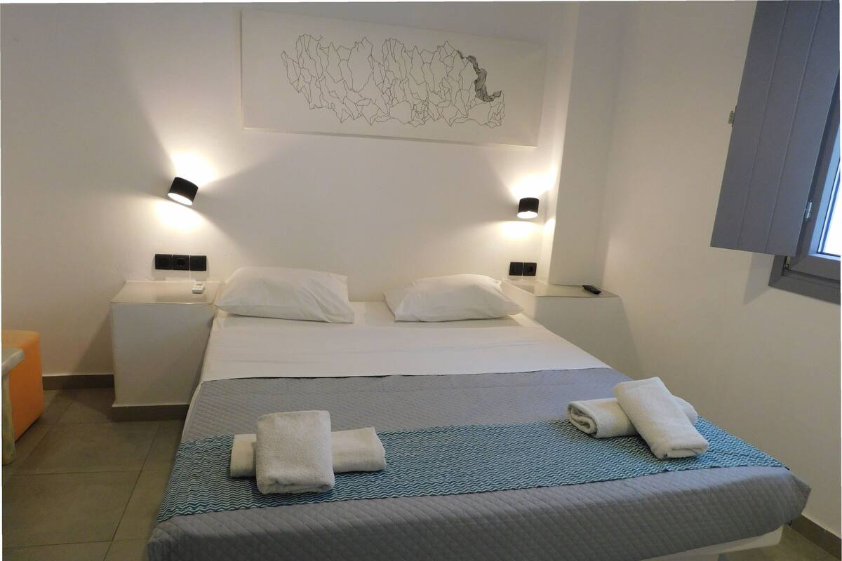 Un lit bien fait, avec du linge de lit blanc et deux oreillers, une table de chevet avec une télécommande, une carte accrochée au mur et une fenêtre donnant sur un beau paysage.