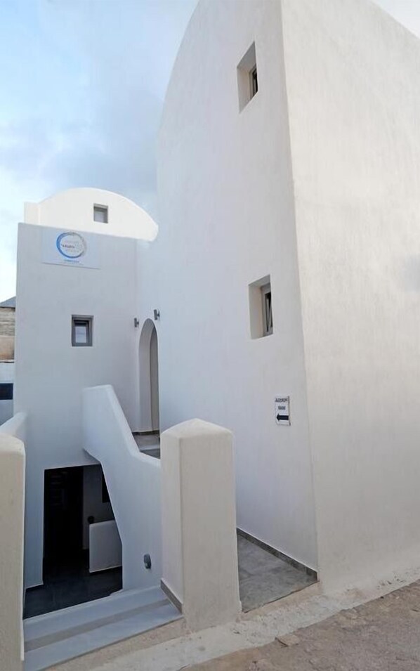 A modern building with a curved facade, a central entrance with steps, and small windows. - Aegean Studios Perissa (Santorini)