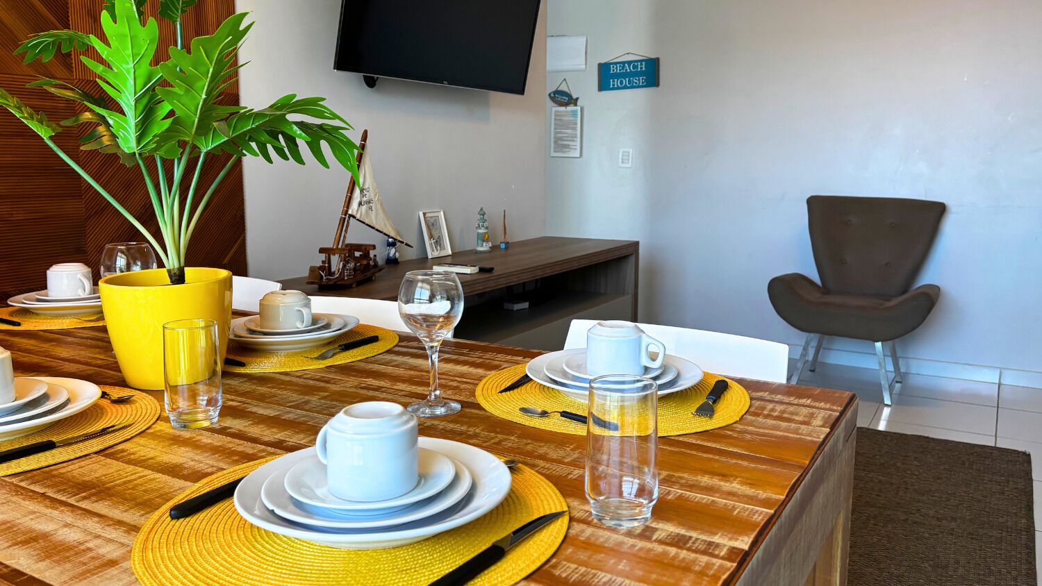 Un coin repas avec une table en bois dressée pour quatre personnes, des sets de table jaunes et une plante en pot.