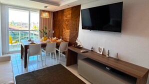 Uma sala de estar moderna com televisão de tela plana na parede, um espaço de jantar com mesa posta para quatro pessoas, e uma grande janela com vista para a cidade.