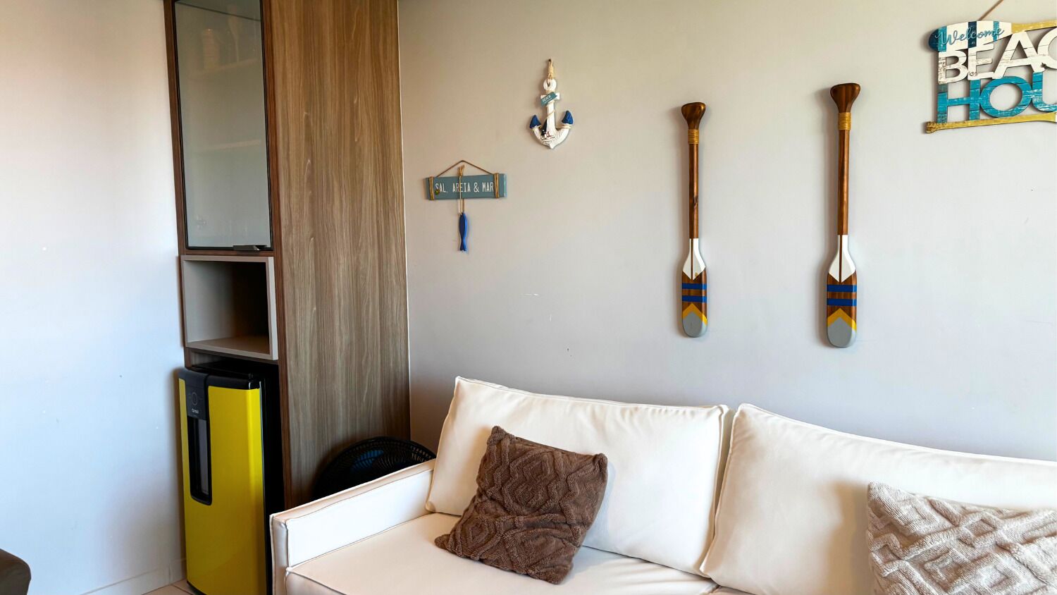 Ein Wohnzimmer mit einem weißen Sofa, einem kleinen gelben Gerät und Wanddekorationen, darunter ein Schild mit der Aufschrift 'SAL AREIA & MAR' und zwei Holzlöffel.