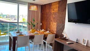Uma área de refeições moderna com uma mesa de madeira posta para quatro pessoas, cadeiras brancas, uma planta em vaso e uma grande janela com vista para a cidade.