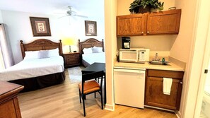 A hotel room with two beds, a desk, a chair, a microwave, a sink, and wooden cabinets.